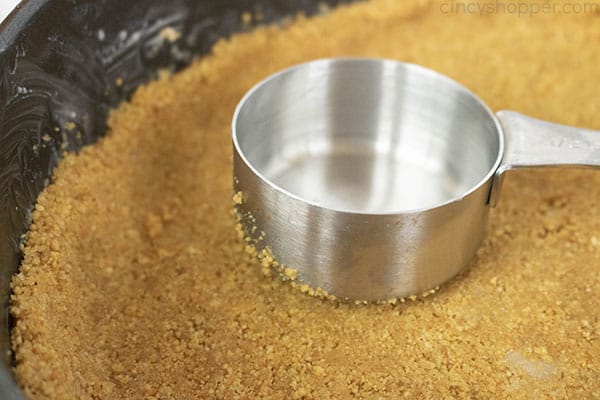 pressing graham cracker crust into a springform pan with a measuring cup