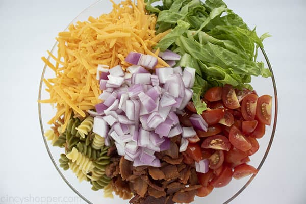 Cooked pasta, shredded cheese, lettuce, tomatoes, bacon, and red onion in a bowl