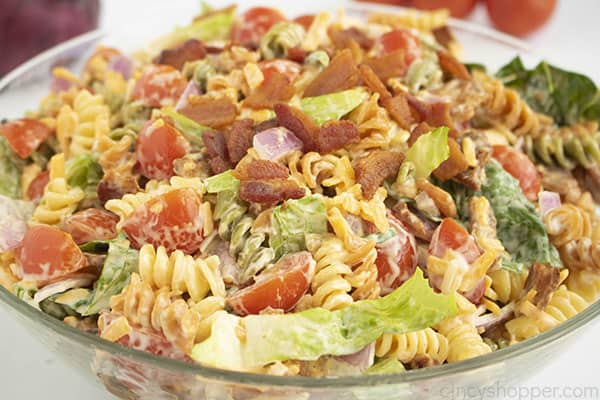 A clear bowl of salad with pasta in front of red onion and tomatoes