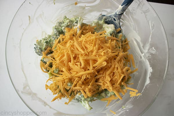 Cheese on top of mixed broccoli in a clear bowl with a blue spatula 