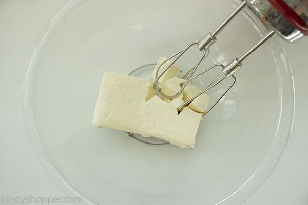 Overhead process shot of cream cheese in a large clear bowl with a hand mixer being used