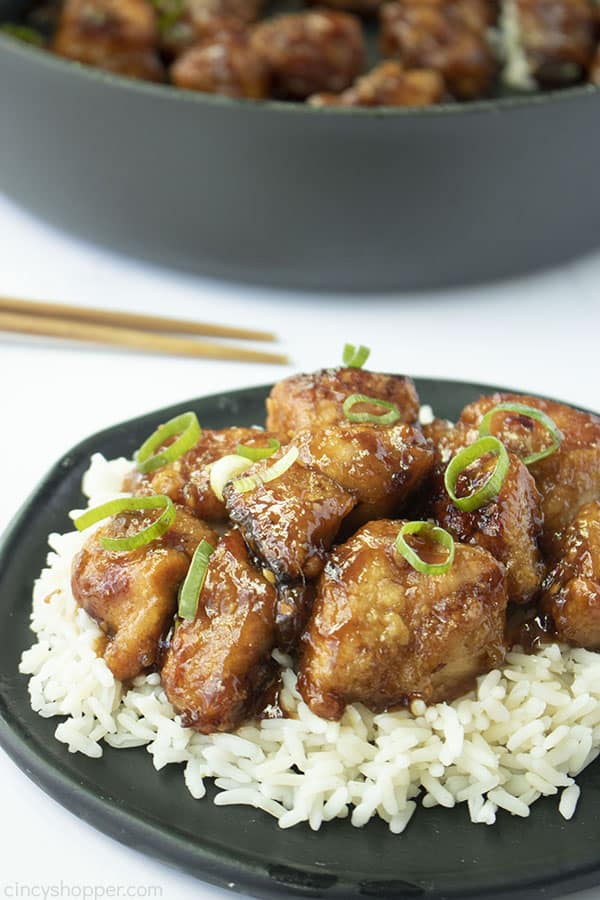General Tso's Chicken on a black plate with rice and pan in back on a white background