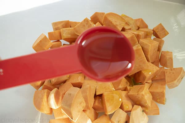 Measuring spoon filled with olive oil pouring on diced sweet potatoes in clear bowl.