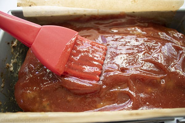 Basting brush with tangy ketchup sauce on top of meatloaf in a pan.