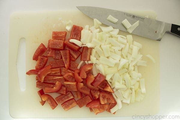 Overhead photo of chopped red peppers and onions on a white cutting board with a large silver knife at the top