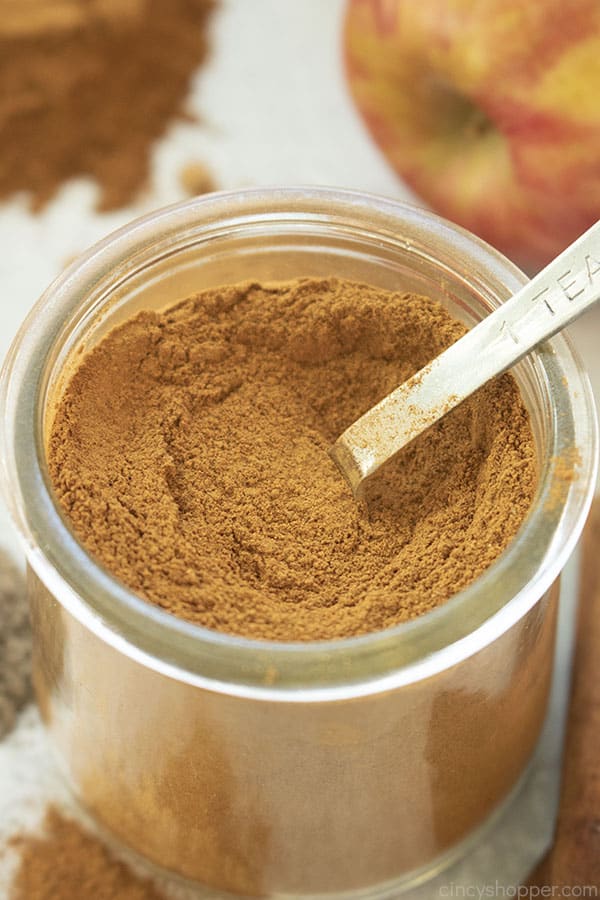 Overhead closeup of Apple Pie Spice mix in a clear jar with spoon. Spices and apple in the background