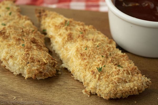 Horizontal image of crispy baked chicken tenders on a cutting board, bbq sauce and red and white napkin in background
