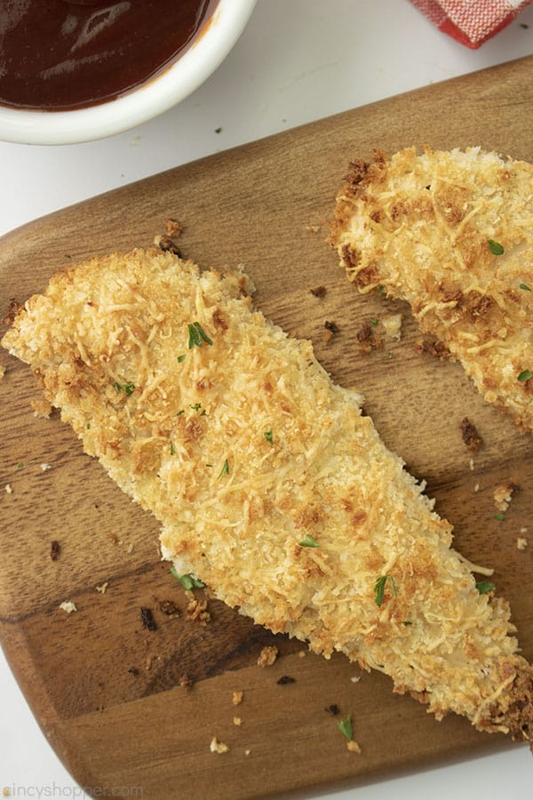 Baked tenders of chicken on a dark cutting board white background and white dish with BBQ sauce