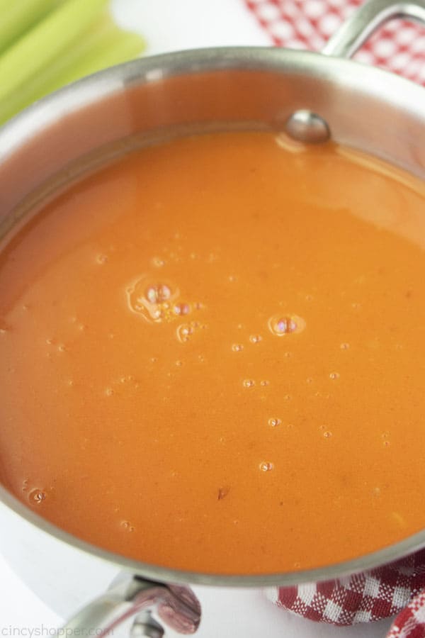 Buffalo Sauce in a stainless pan with red and white checkered towel, celery and white background