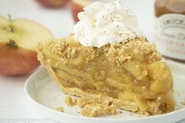 Finished apple pie slice with whipped cream. Apples and caramel in the background.