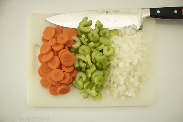 Chopped carrots, celery and onions on a white cutting board with a knife