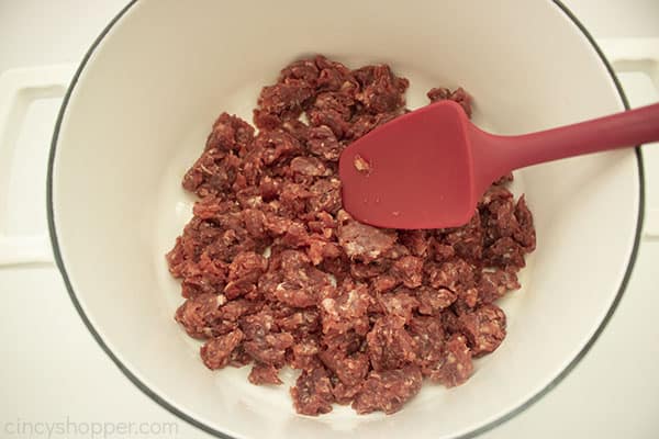 Raw ground beef added to white pot with a red spatula