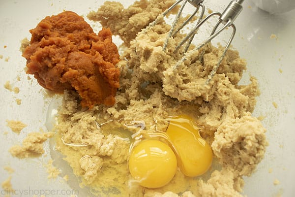 Eggs and pumpkin added to blended sugar mixture in a clear bowl with beaters