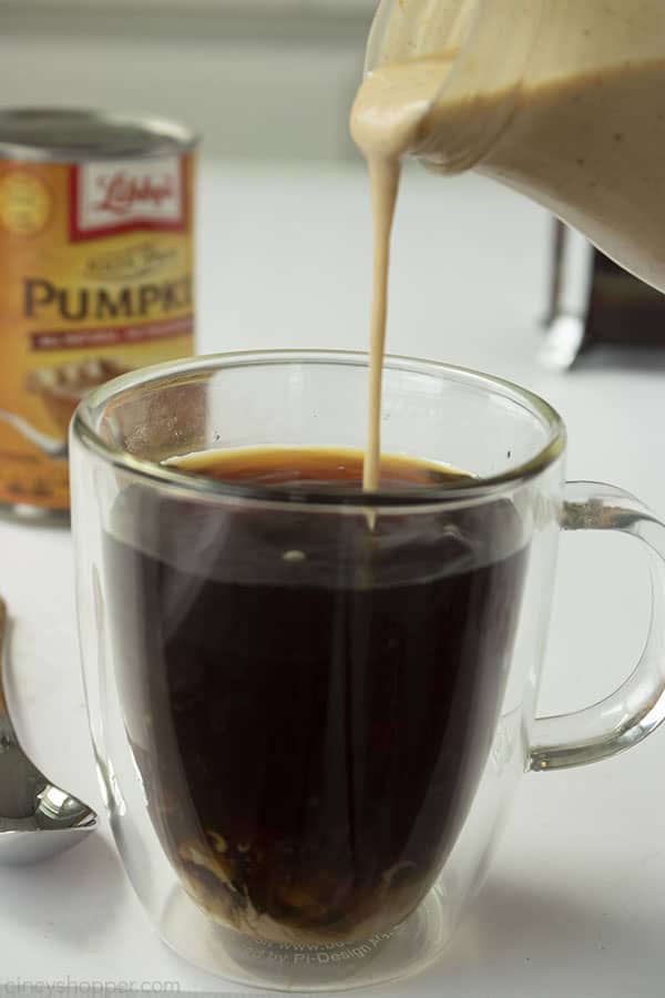 Clear coffee mug with homemade pumpkin creamer being poured in. White background with canned pumpkin in the distance.