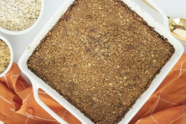 Horizontal image of baked crisp in a white dish with orange napkin, oats, brown sugar and gold spoon.