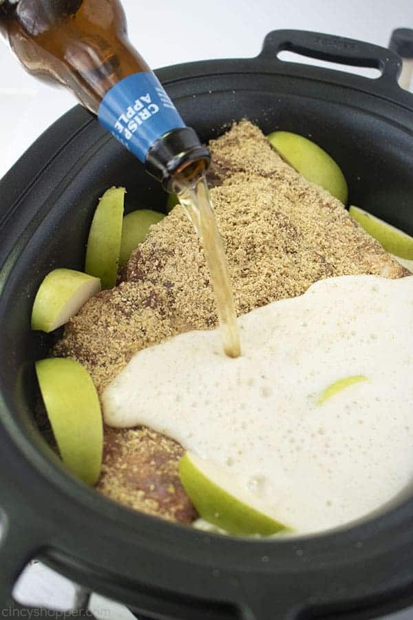 Overhead image of the Pulled Pork in a black oval Slow Cooker topped with the apple crisp rub, sliced green apples, and crisp apple cider being poured over 