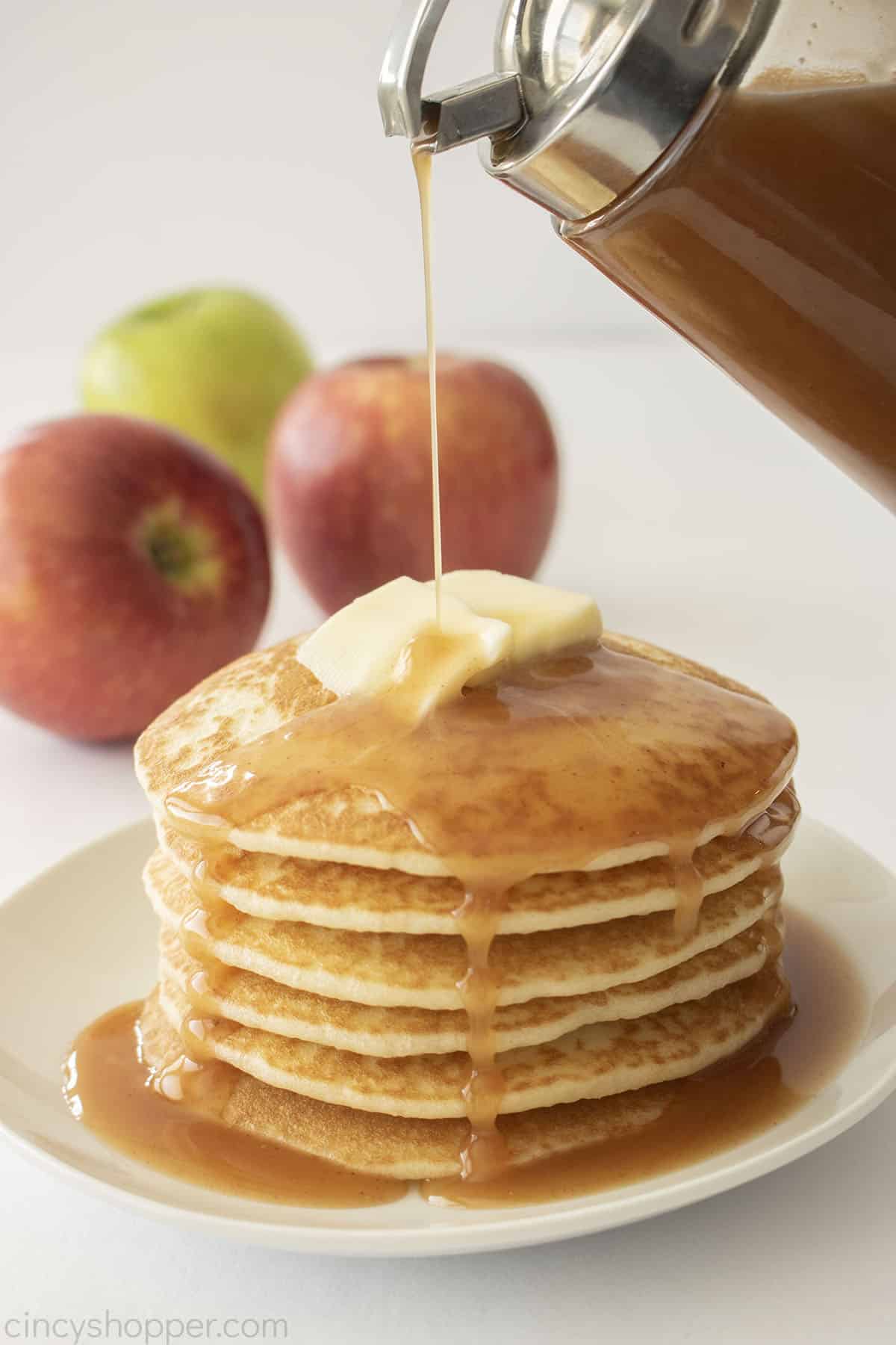 Stack of pancakes with syrup pouring on top- apples in the background.