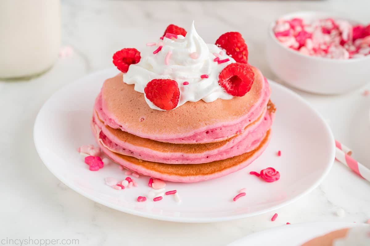 Plate with pink colored pancakes with hipped topping, sprinkles and fruit.