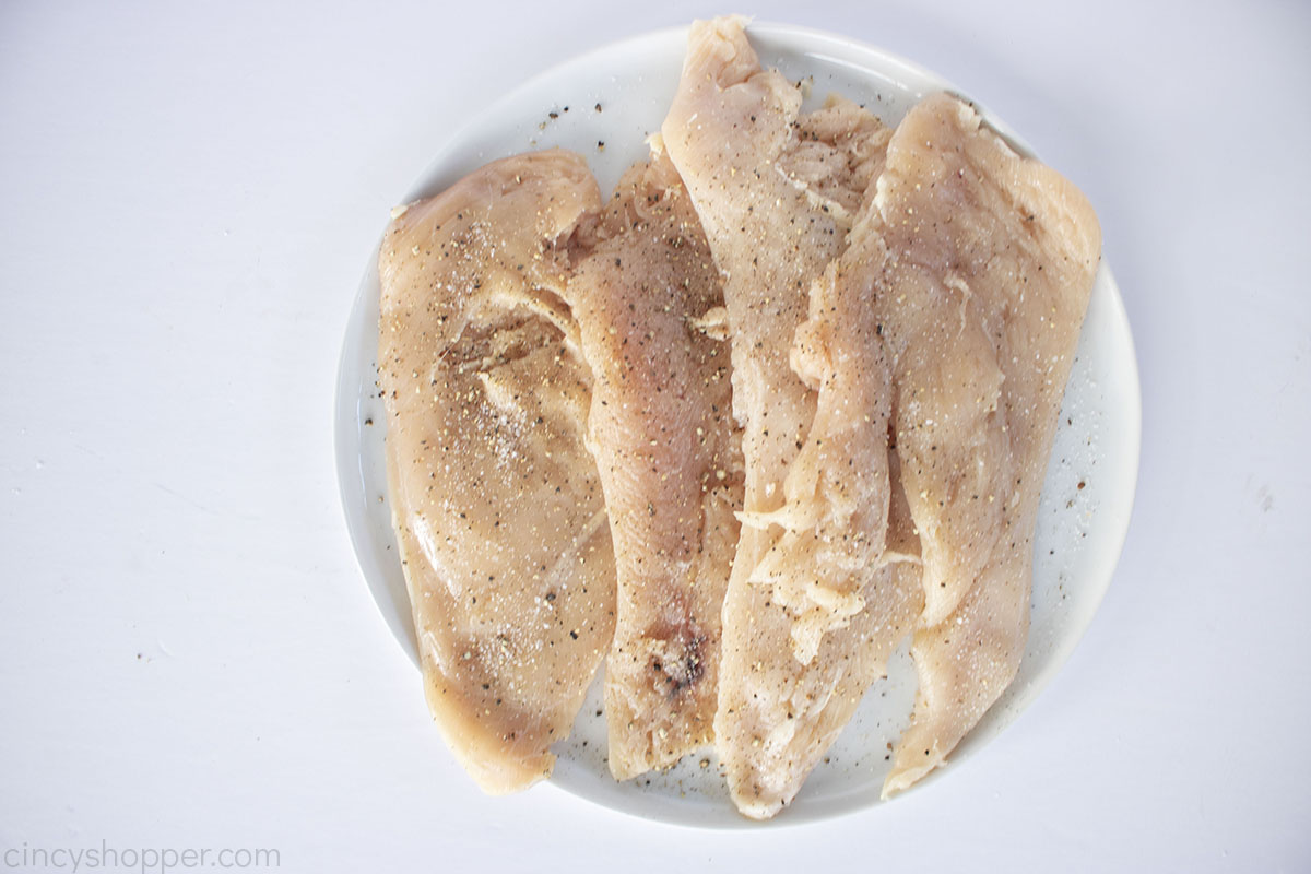 Raw chicken breasts butterflied and pounded with salt and pepper on a white plate.
