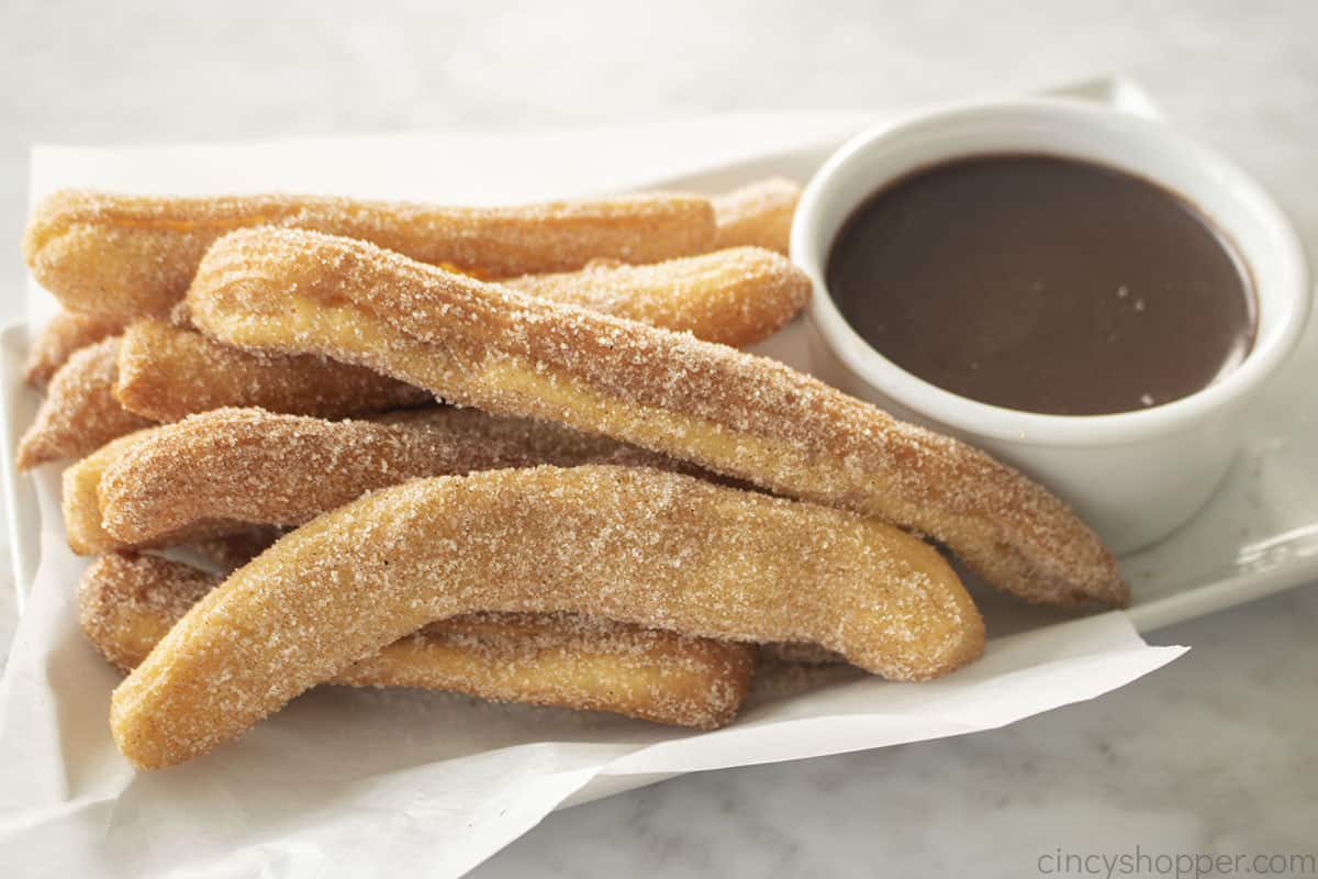 Easy Authentic Churros on a white platter with chocolate dipping sauce.