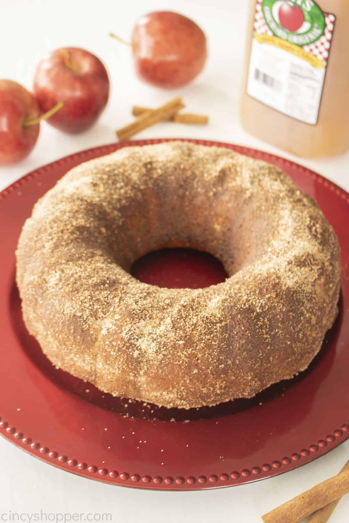 Apple Cider Donut Cake on a red plate with cinnamon sticks, apples and cider.