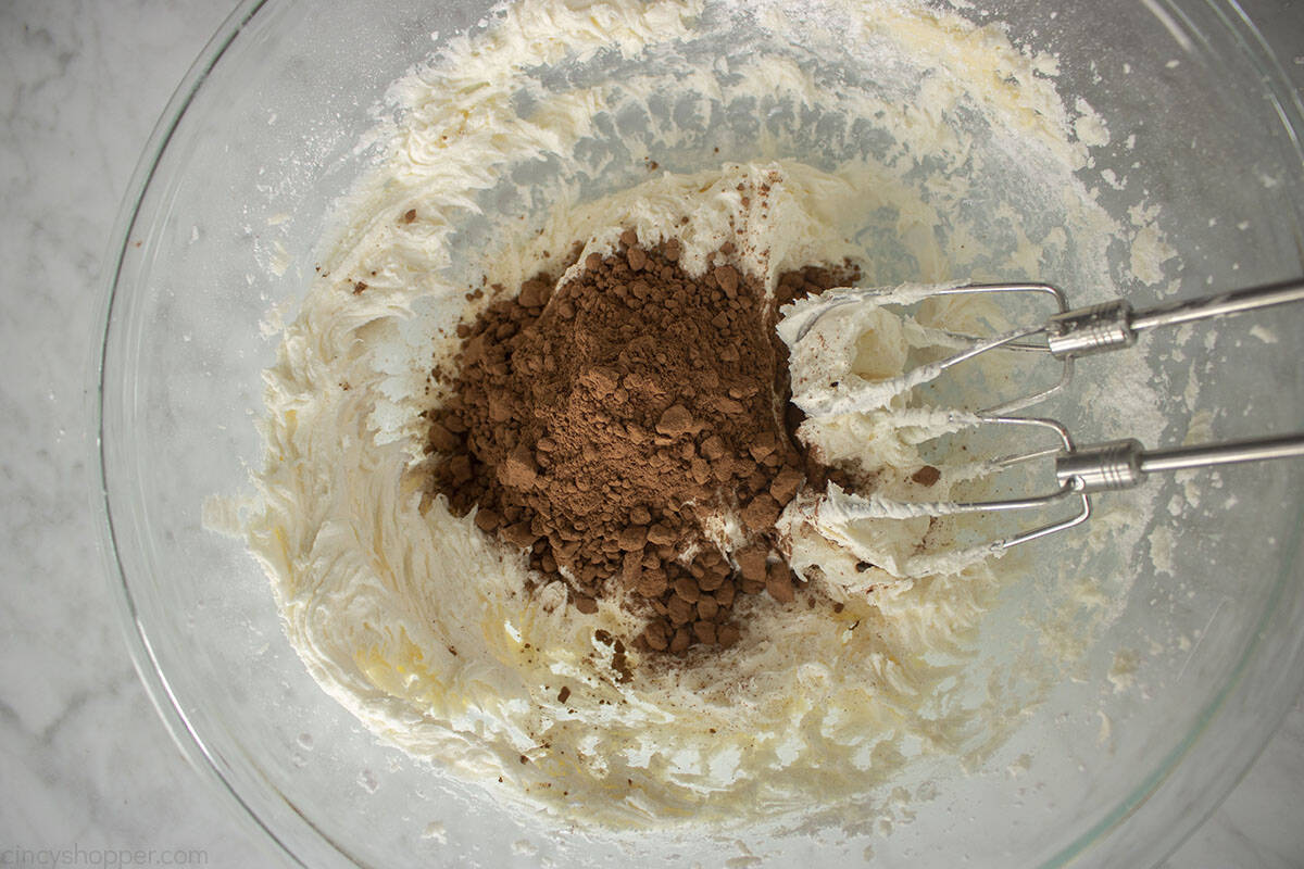 Cocoa added to butter cream frosting in a bowl with hand mixer.