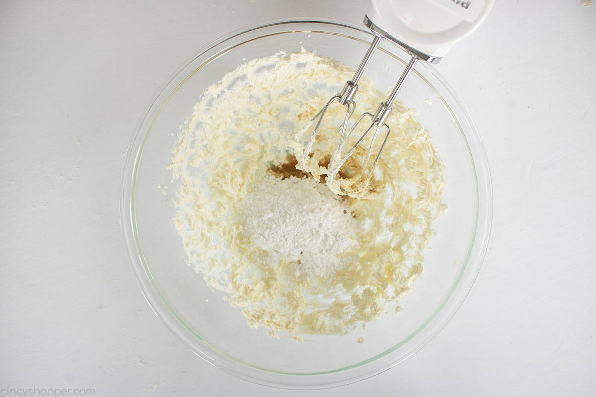 Cream cheese, powdered sugar and vanilla in a clear bowl with hand mixer.