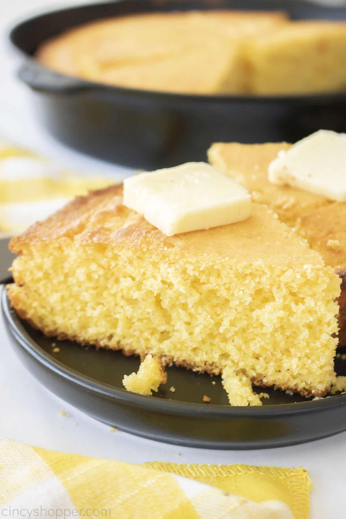 Slice of from scratch. cornbread on a black plate with pat of butter.