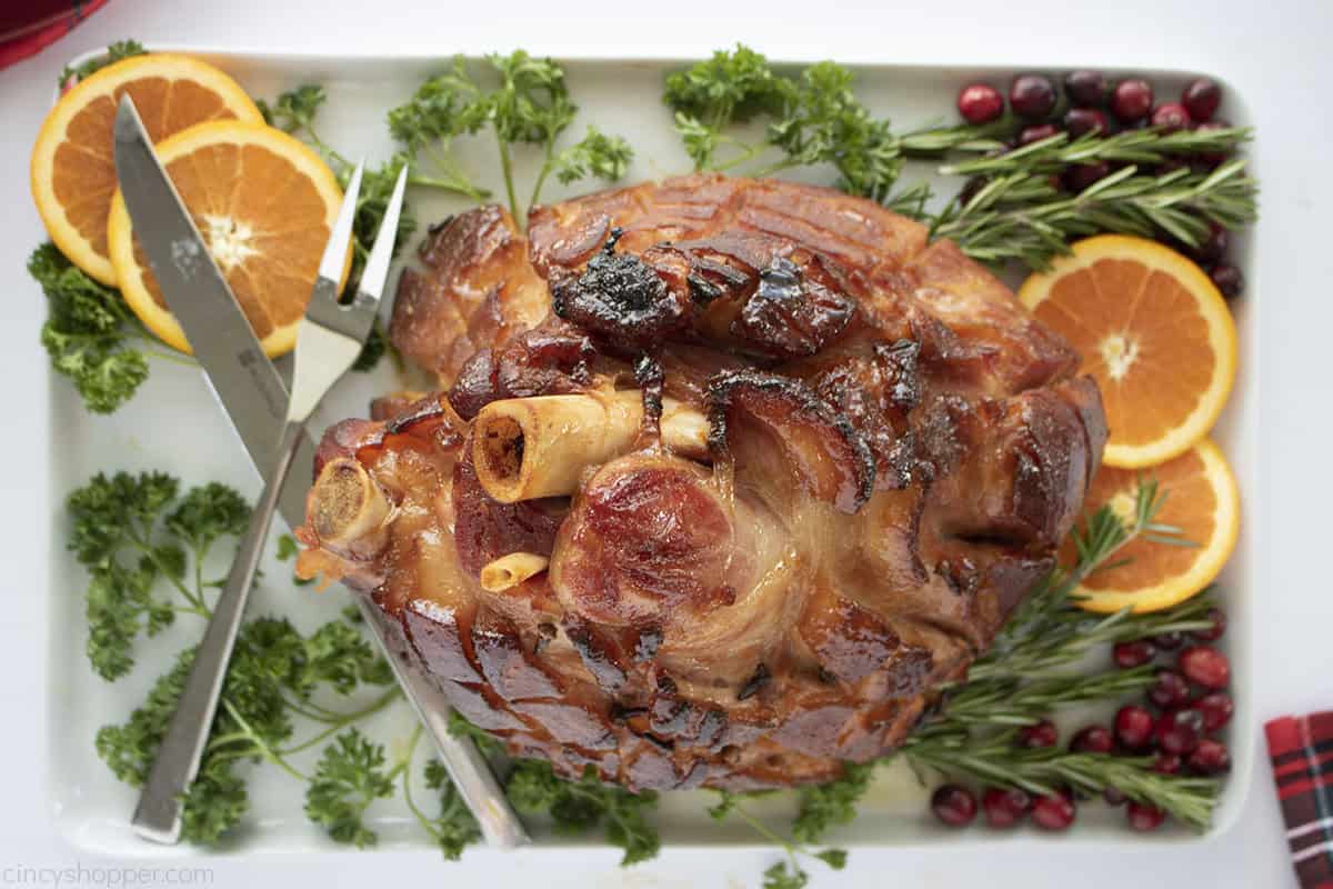 Overhead sideways view of baked ham with bourbon glaze on a platter with oranges and fresh herbs.