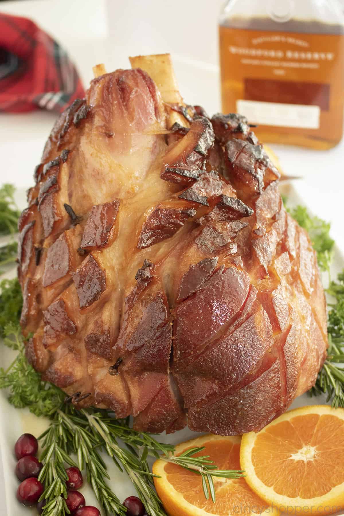 Whole glazed ham on a platter uncut with fresh herbs.