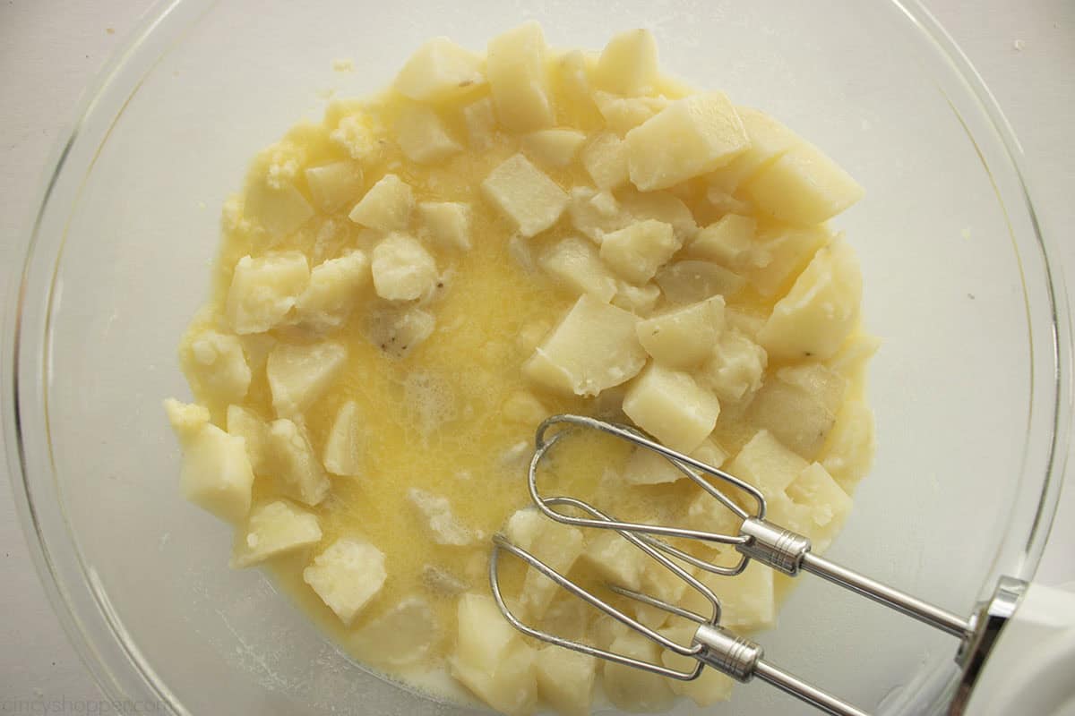 Some milk and butter mixture added to cooked potatoes in a bowl with hand mixer.
