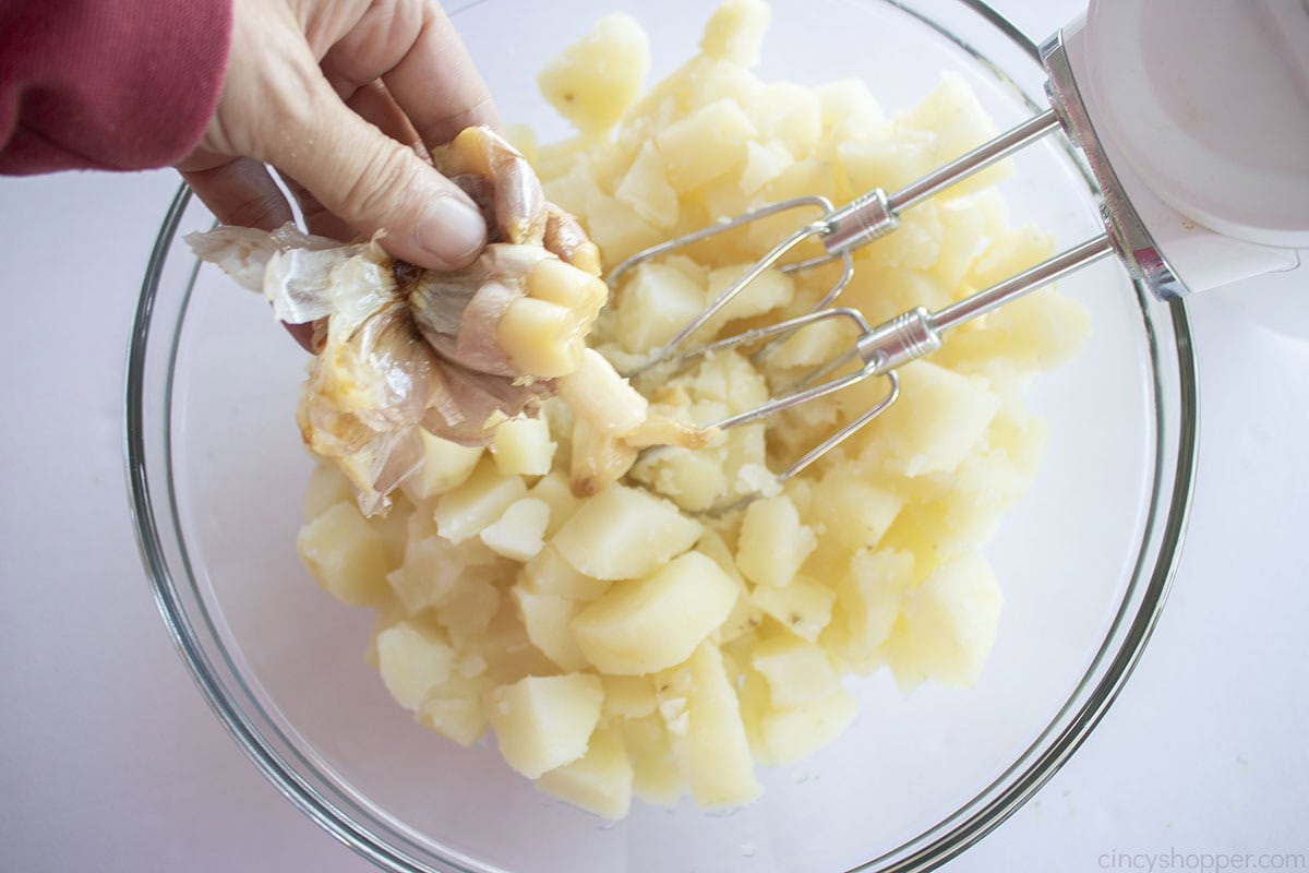 Smooshed garlic being added to boiled potatoes in a mixing bowl with hand mixer.