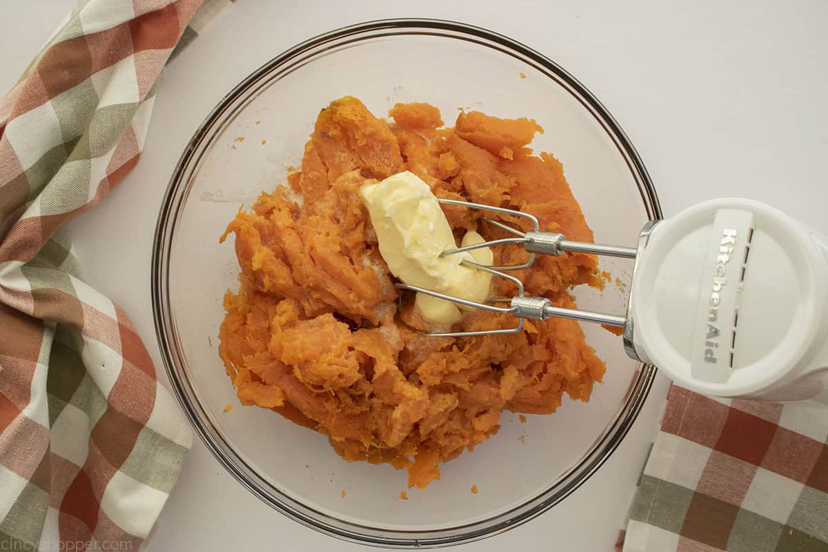 Cooked sweet potatoes in a clear bowl with butter and a hand mixer.