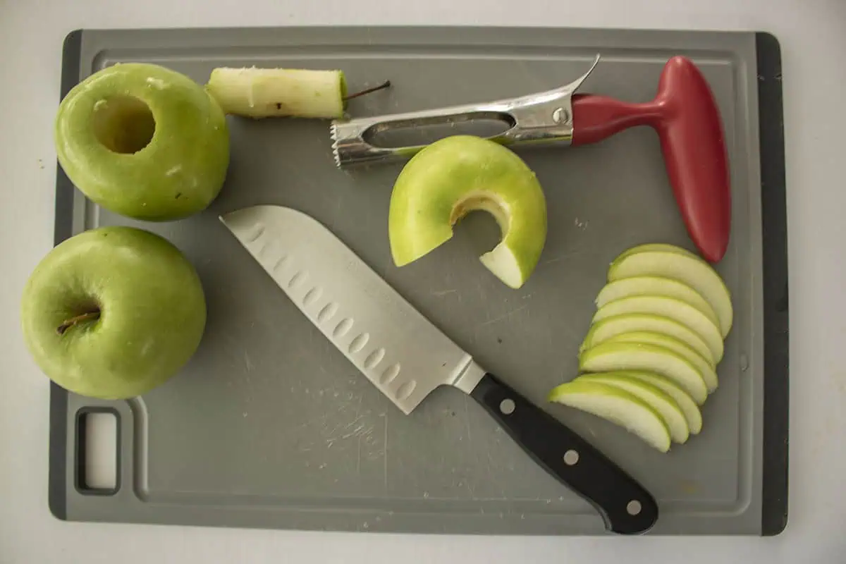Cored and sliced apples on a cutting board with knife and apple corer.
